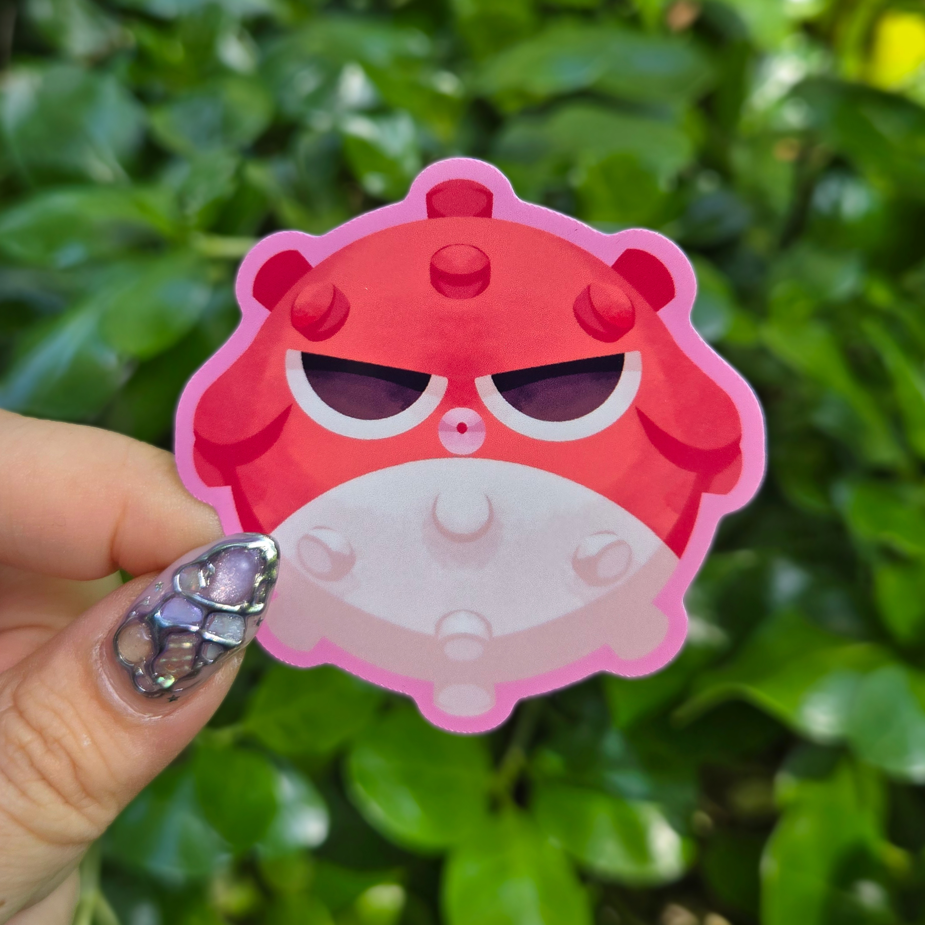 Sticker design on the centre of the image held up with fingers against a green shrubbery background