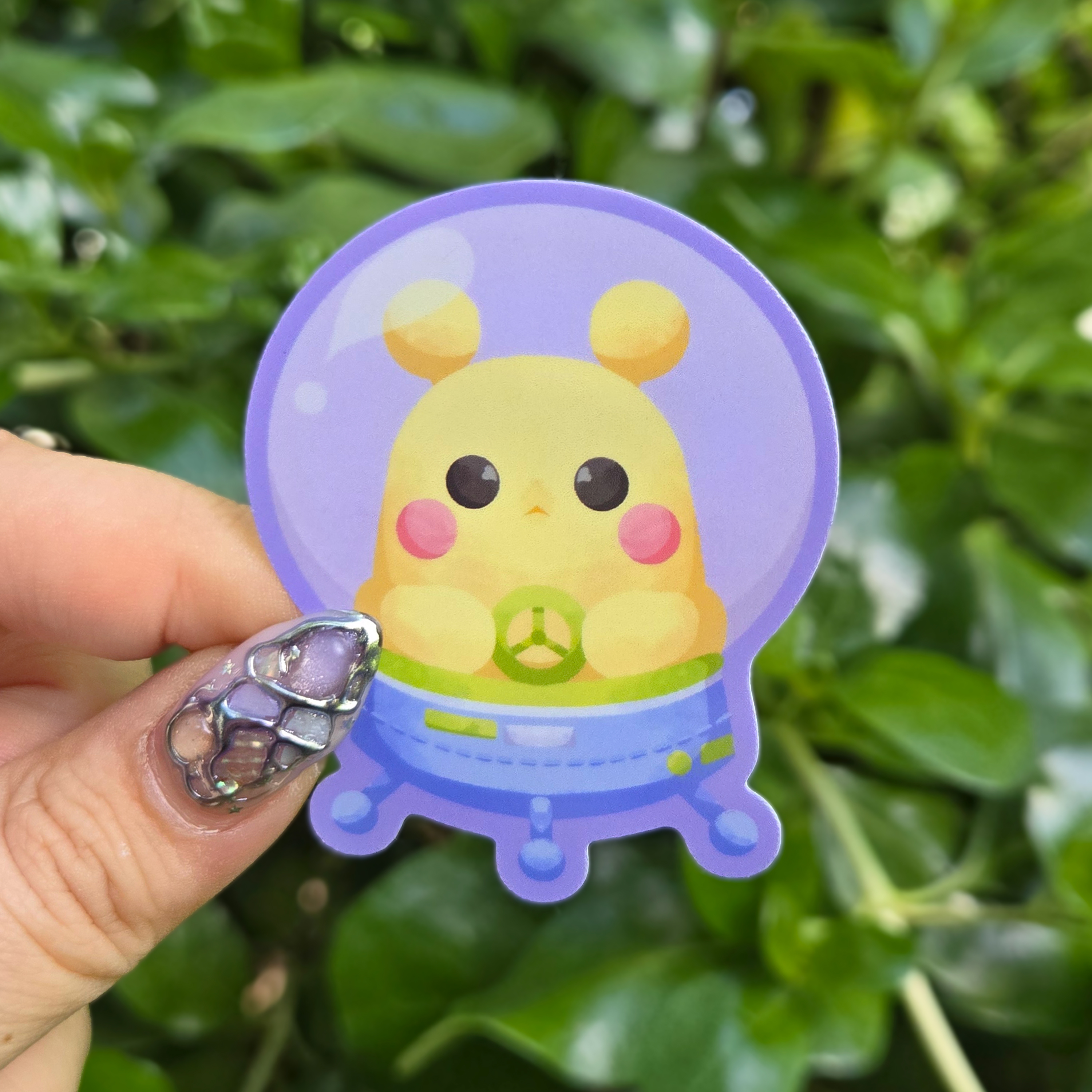 Sticker design on the centre of the image held up with fingers against a green shrubbery background