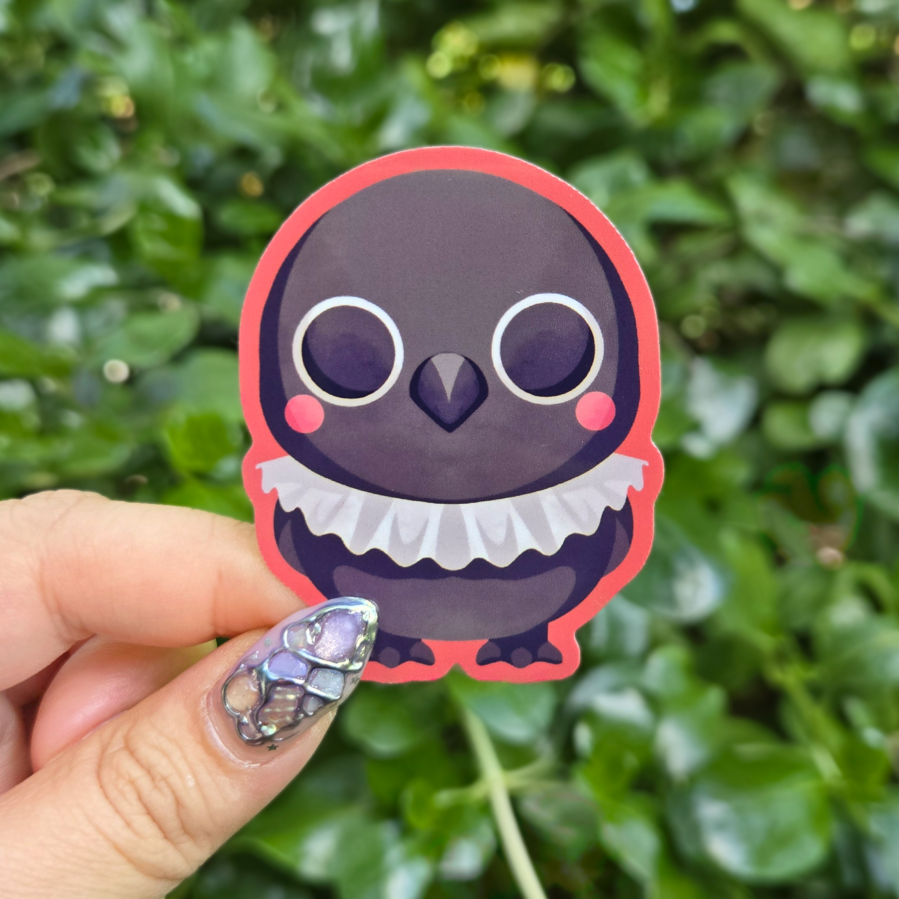 Sticker design on the centre of the image held up with fingers against a green shrubbery background