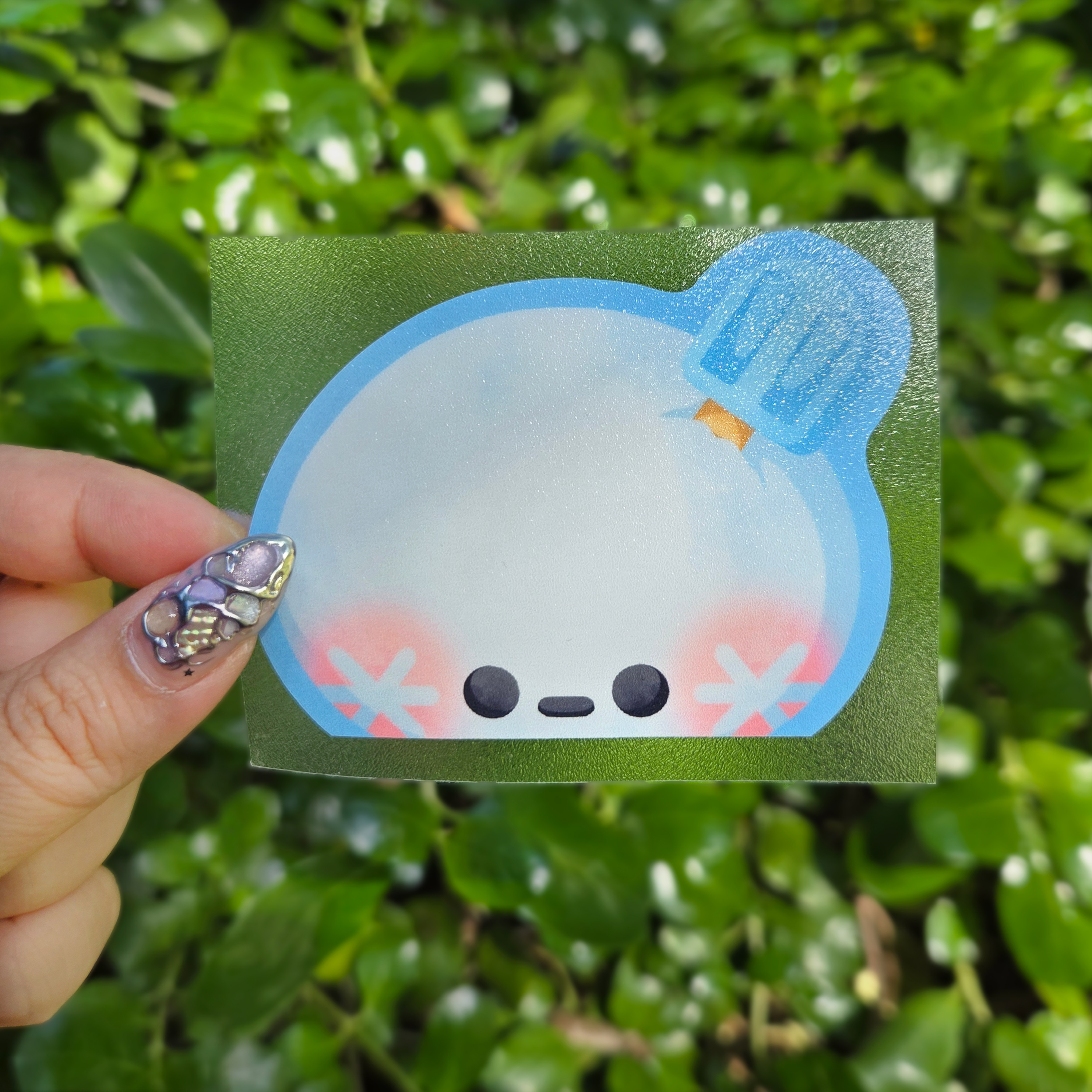 Peeker design held up with a clear transfer tape at the centre of the image by fingers. Green shrubbery background
