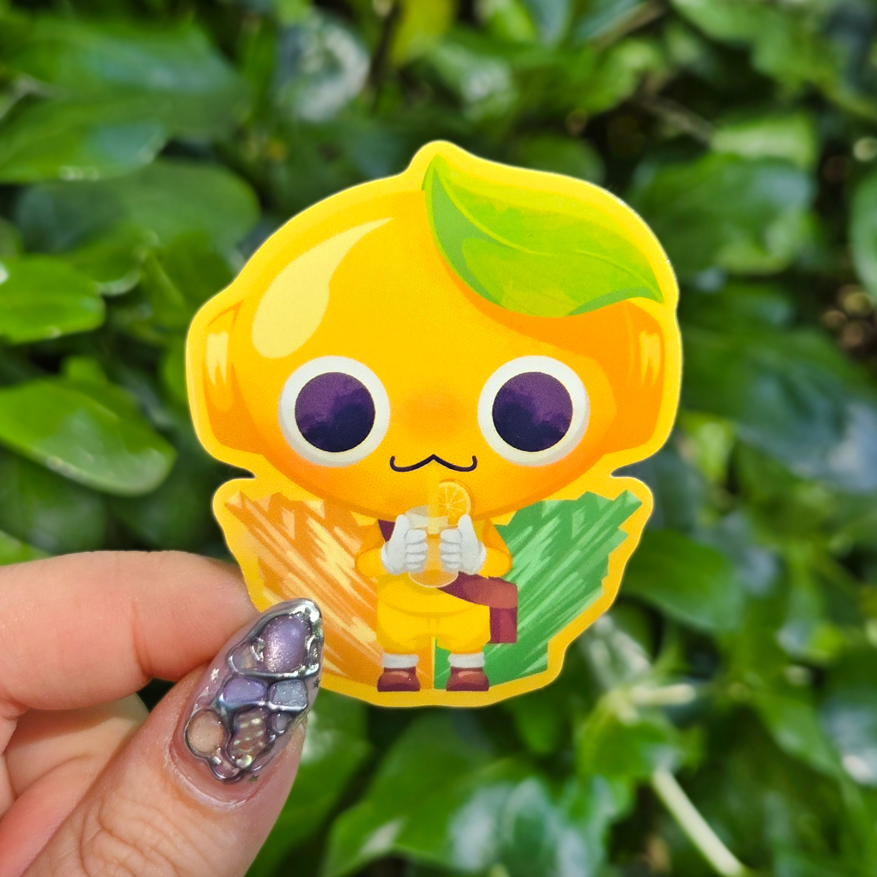 Sticker design on the centre of the image held up with fingers against a green shrubbery background