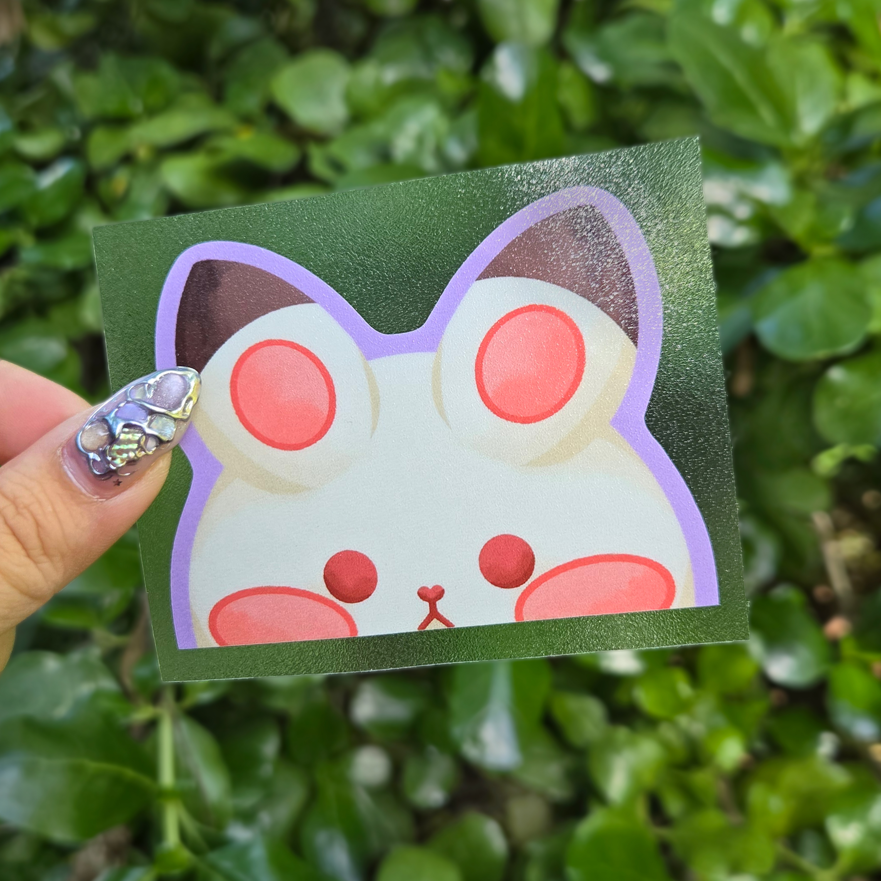 Peeker design held up with a clear transfer tape at the centre of the image by fingers. Green shrubbery background