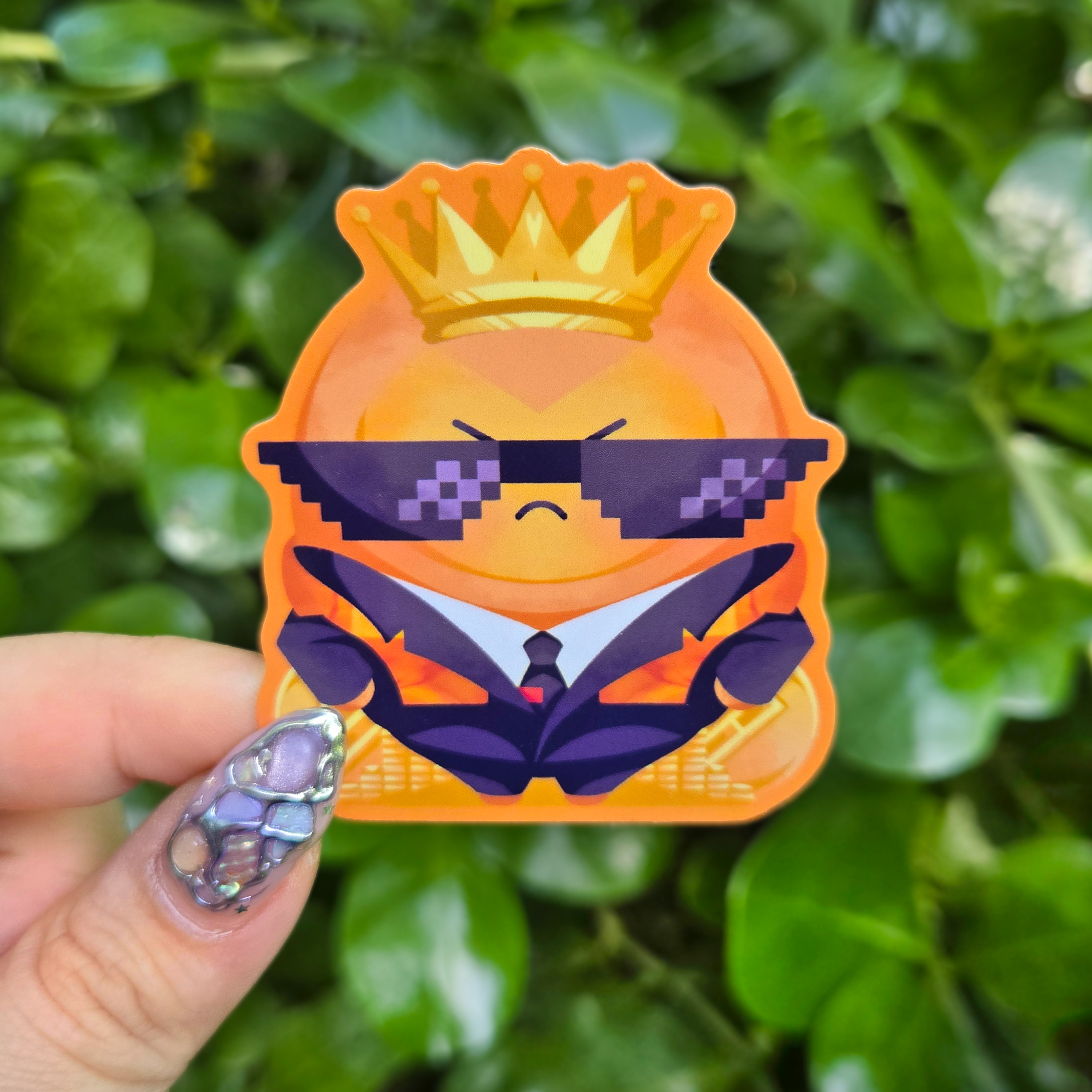 Sticker design on the centre of the image held up with fingers against a green shrubbery background