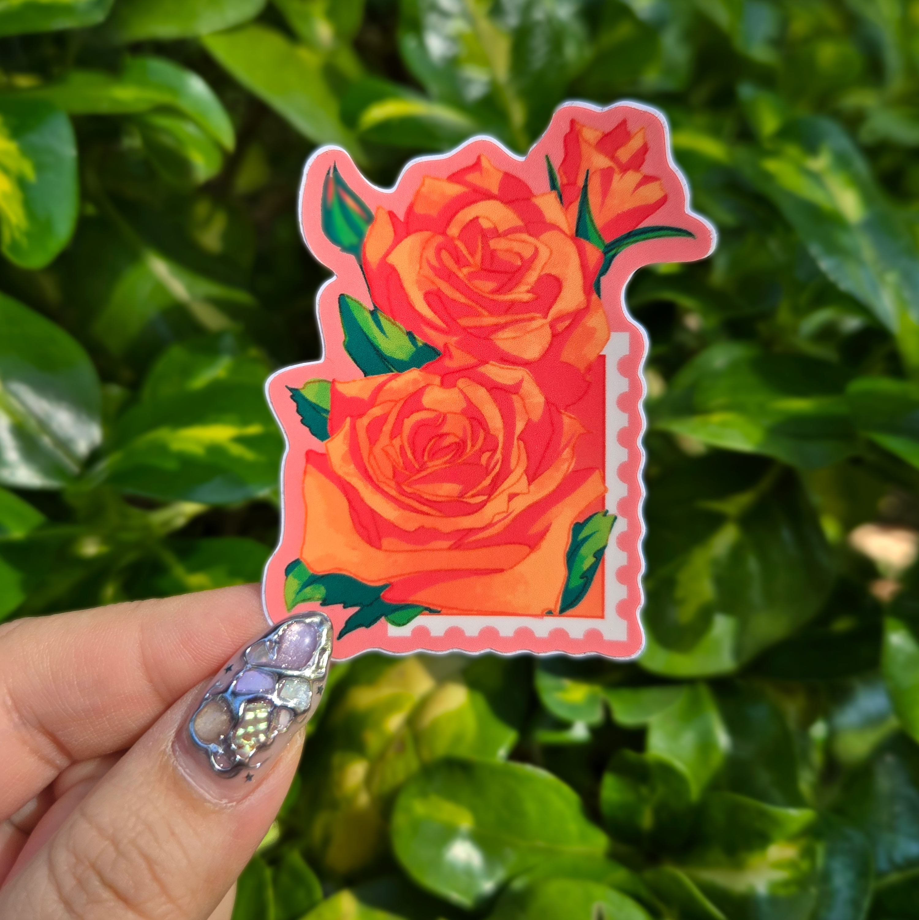 Sticker design on the centre of the image held up with fingers against a green shrubbery background