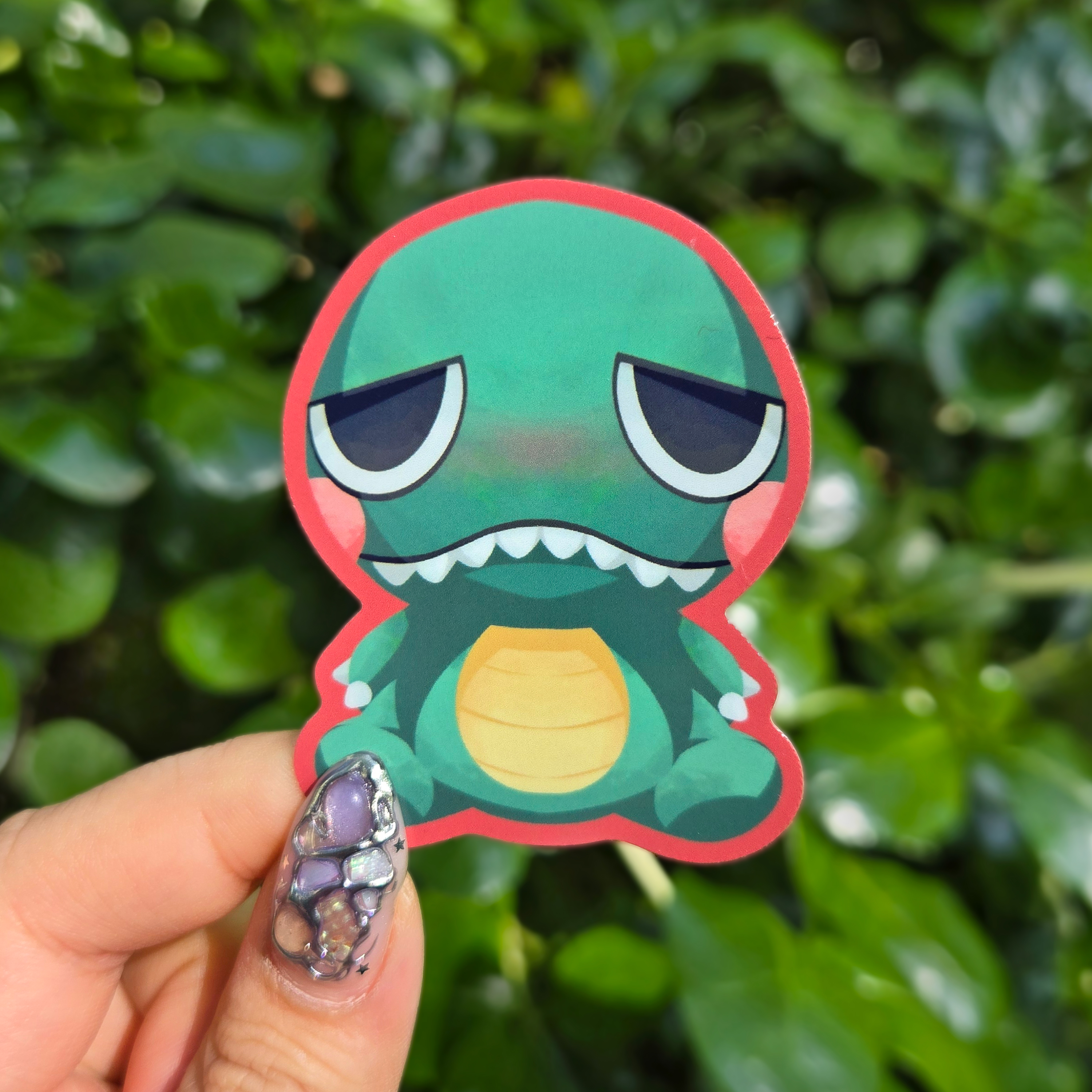 Sticker design on the centre of the image held up with fingers against a green shrubbery background