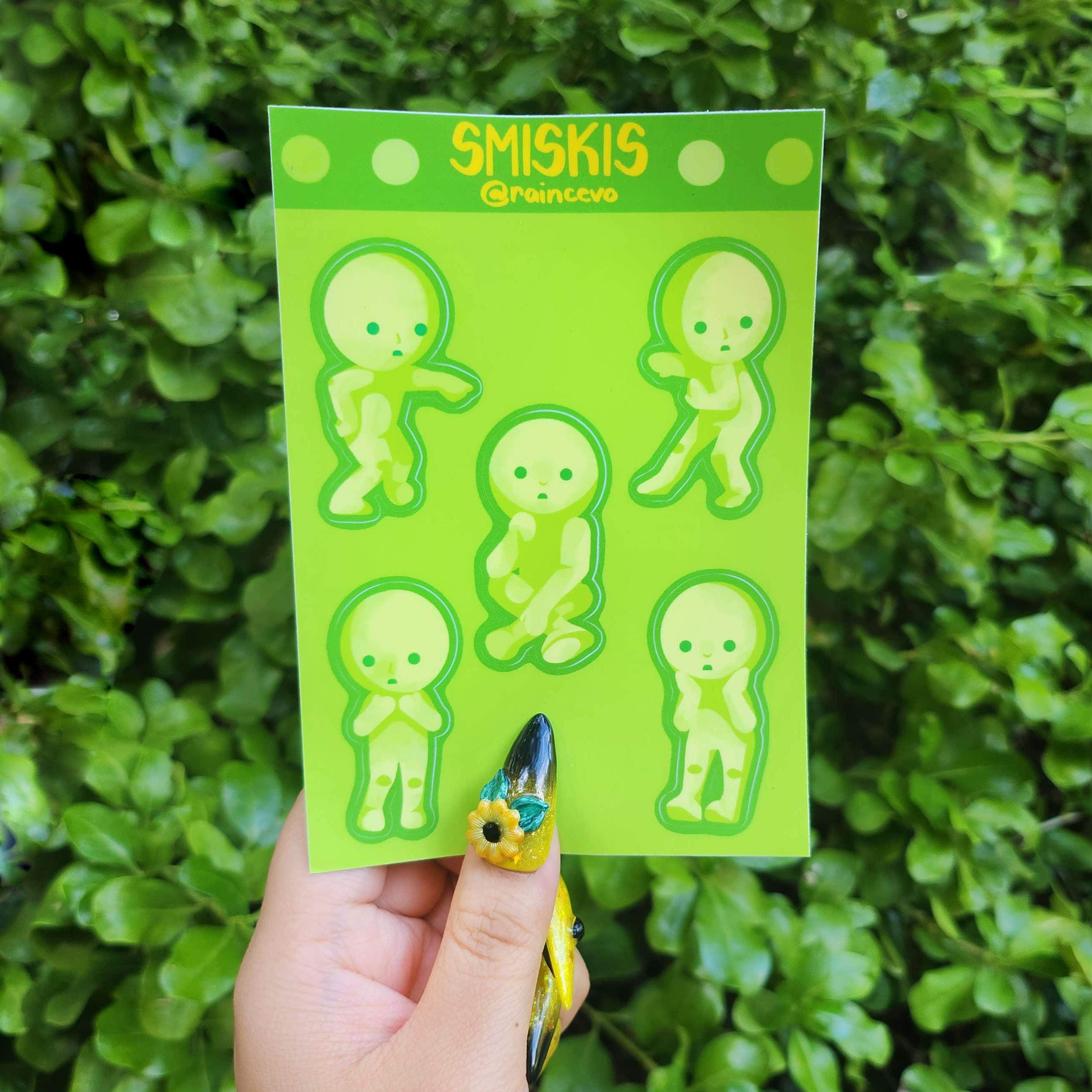 Hand holding up sticker sheet against green shrubbery background. Sticker sheet includes designs of Smiskis in different poses.