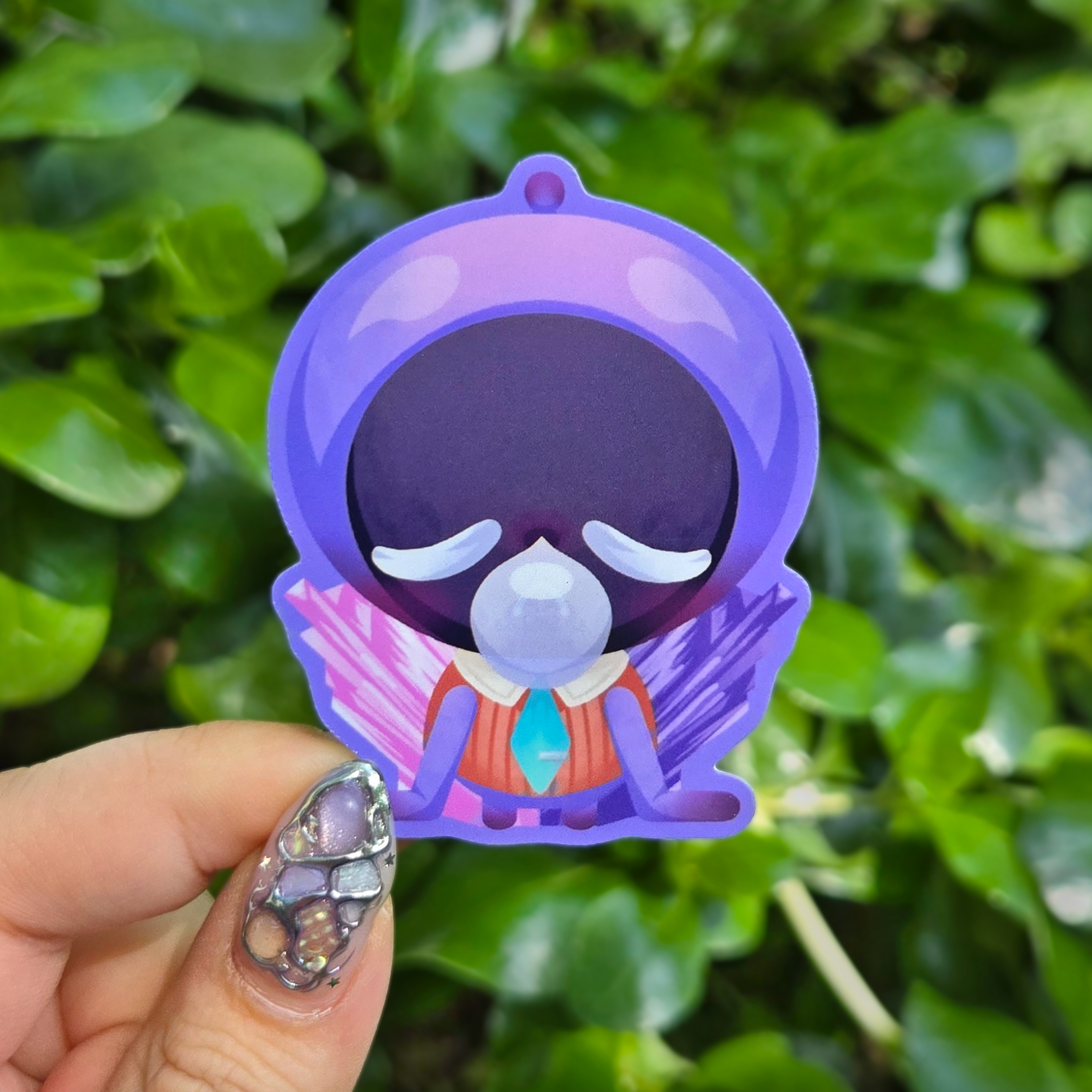 Sticker design on the centre of the image held up with fingers against a green shrubbery background