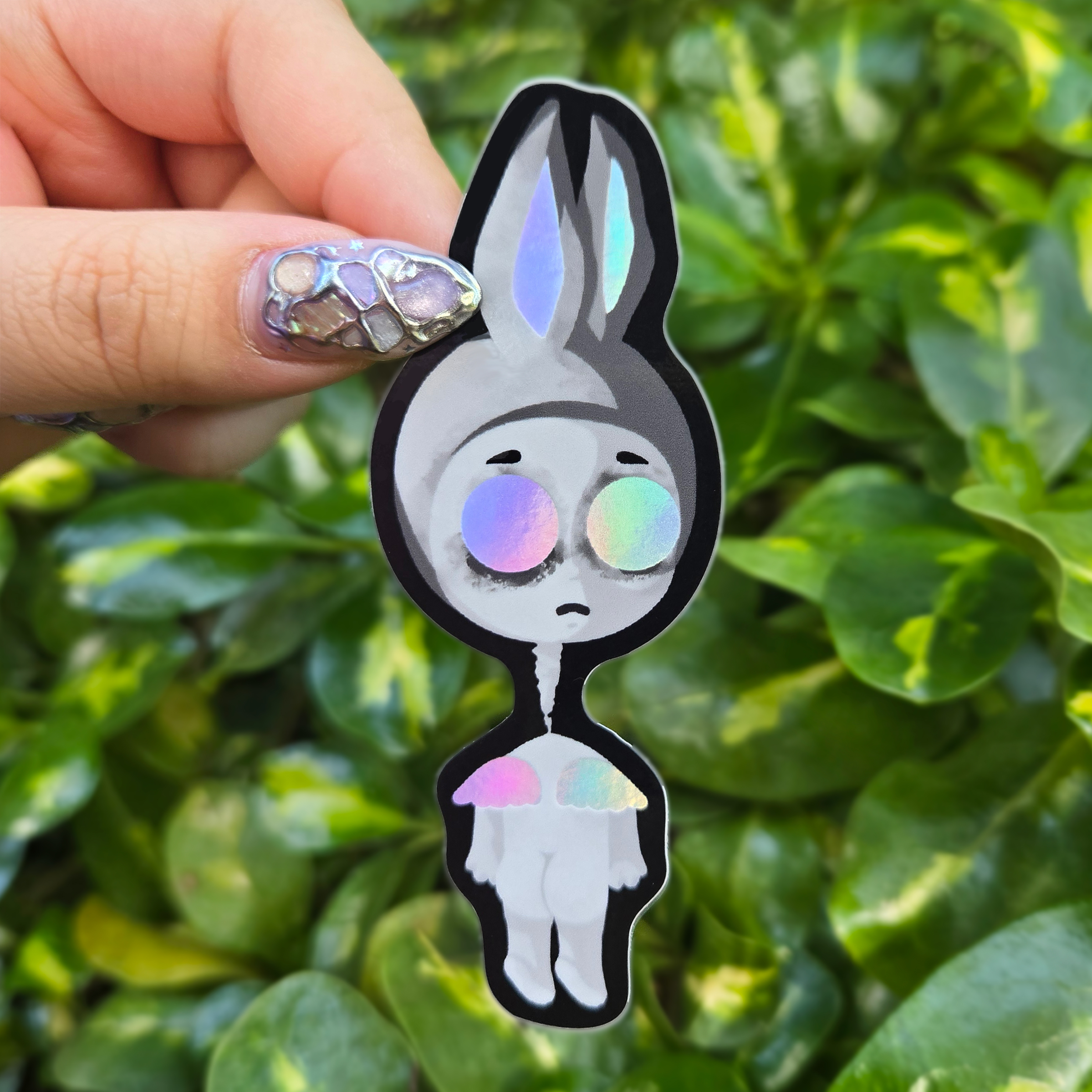 Sticker design with spot holo held up by fingers against green shrubbery background.