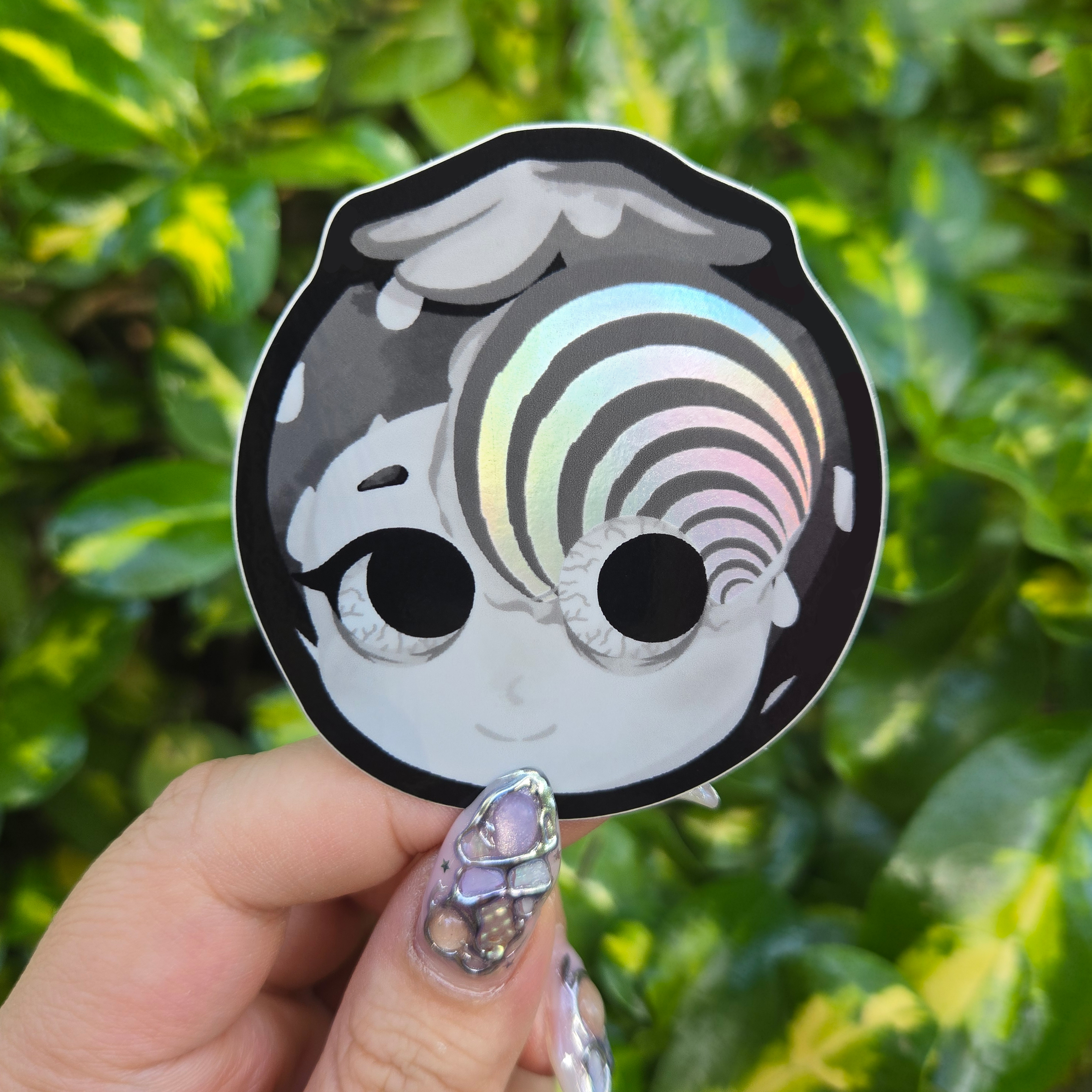 Sticker design with spot holo held up by fingers against green shrubbery background.