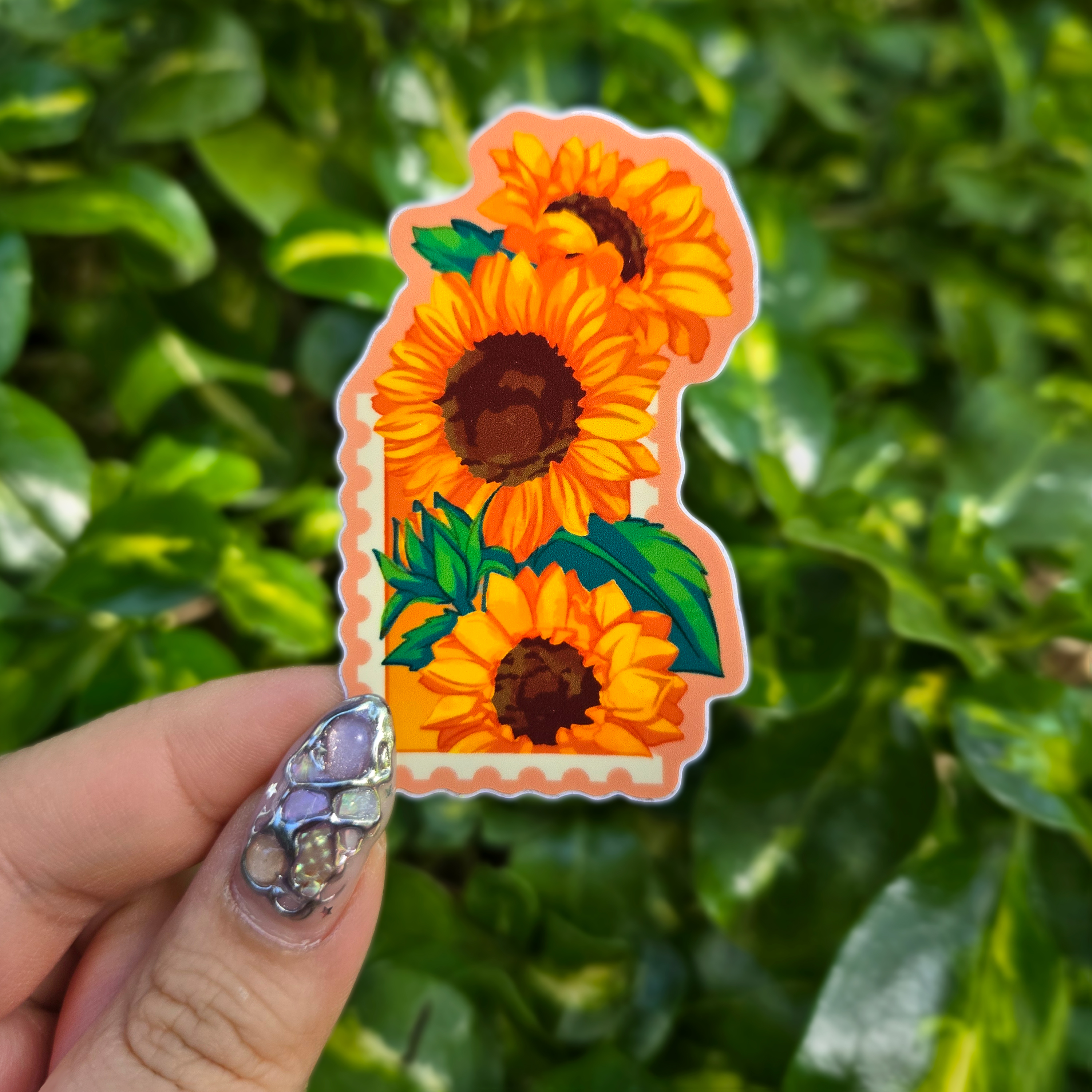 Sticker design on the centre of the image held up with fingers against a green shrubbery background