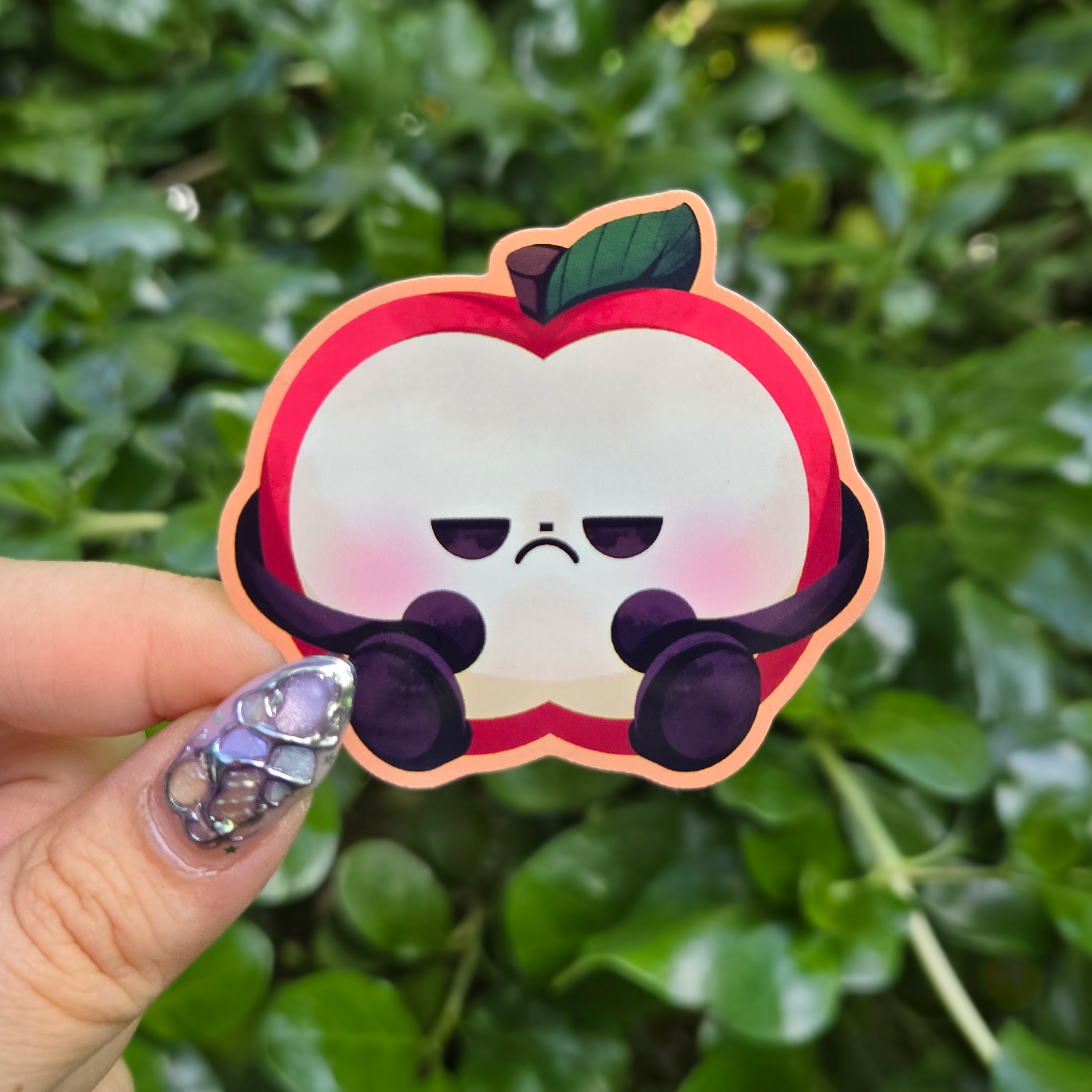 Sticker design on the centre of the image held up with fingers against a green shrubbery background