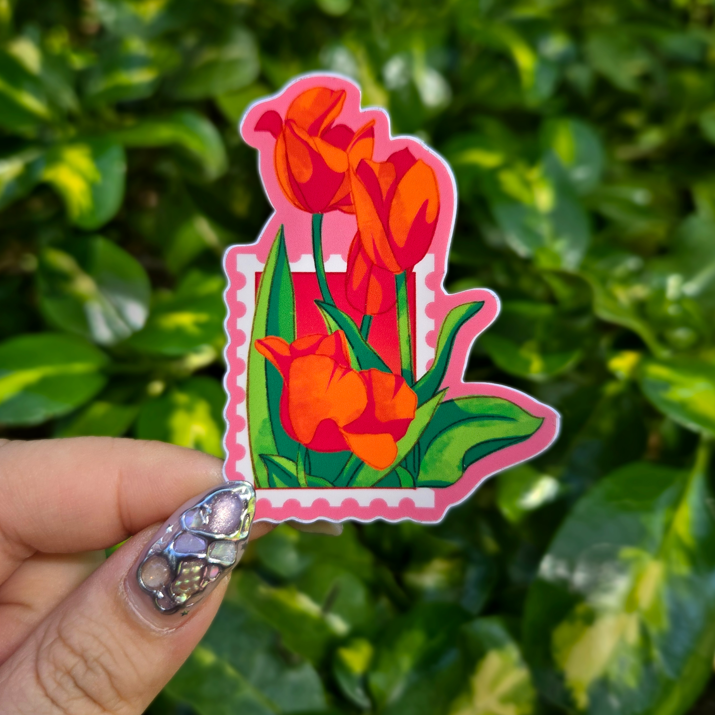 Sticker design on the centre of the image held up with fingers against a green shrubbery background